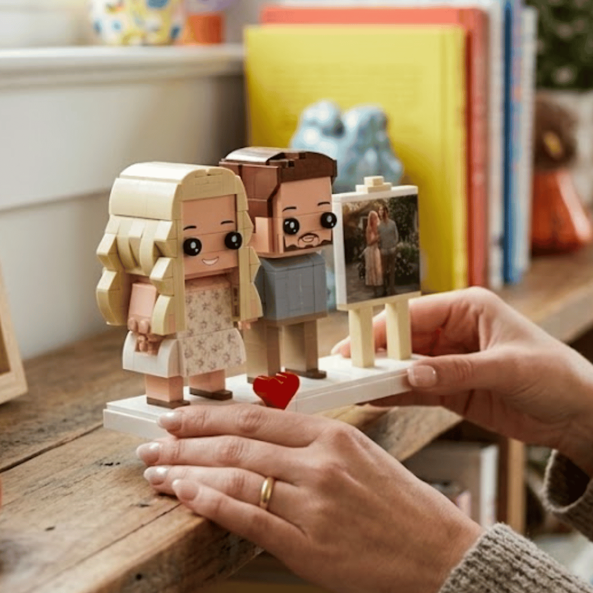 A person's hands holding a custom couple brick figure photo on a rustic wooden table, with colorful books in the background.