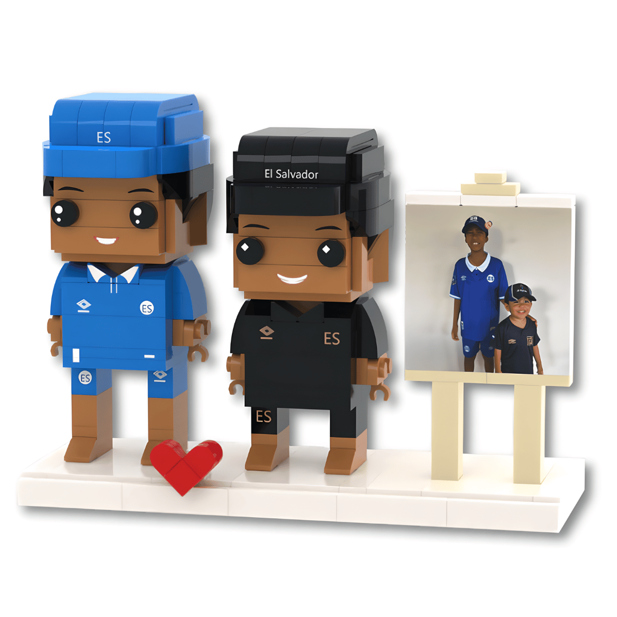 Two custom Lego-style figurines representing soccer fans, one in a blue Chelsea kit and one in a black El Salvador national team kit, standing on a white base next to a small easel displaying a reference photo. A red heart icon is placed in front of the blue figure.
