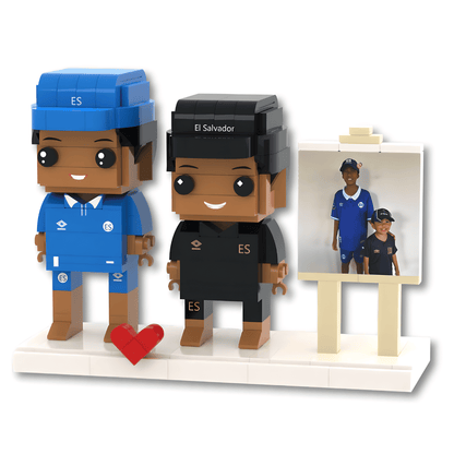 Two custom Lego-style figurines representing soccer fans, one in a blue Chelsea kit and one in a black El Salvador national team kit, standing on a white base next to a small easel displaying a reference photo. A red heart icon is placed in front of the blue figure.
