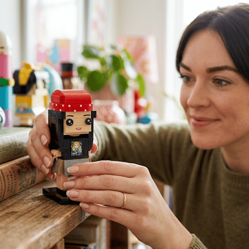 A woman in an olive green sweater holds a small LEGO minifigure with a red bandana and black outfit on a wooden shelf in a brightly lit room.