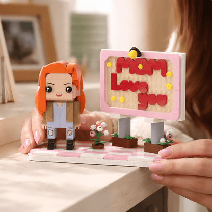 Toy figure of a person with orange hair and a sign saying 'Mom Love You' held by hands.