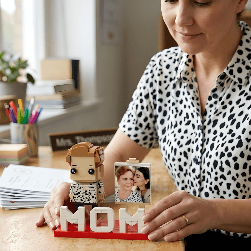 Woman holding a LEGO figure and a photo of two people, with 'MOM' letters on a desk.