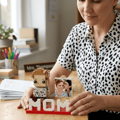 Woman holding a LEGO figure and a photo of two people, with 'MOM' letters on a desk.